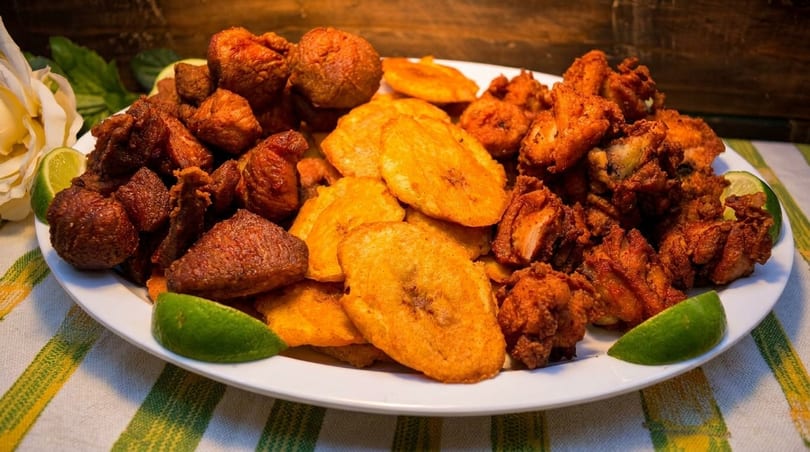 A white plate piled high with fried pork, fried plantains, and fried chicken, garnished with lime wedges.
