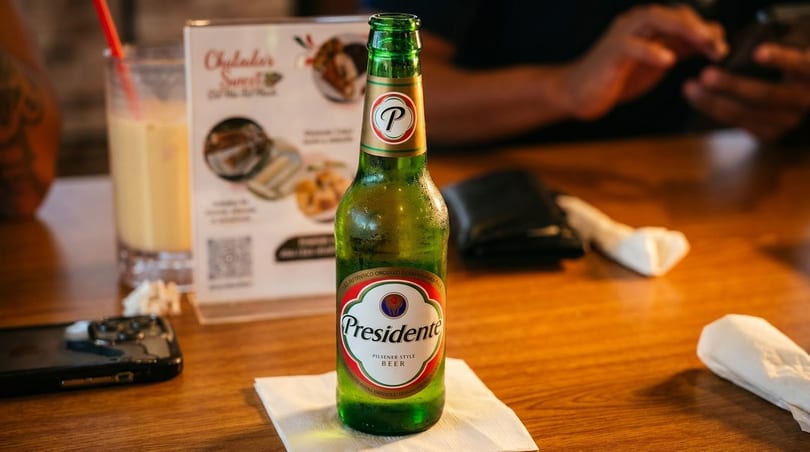 A cold bottle of Presidente beer on a wooden table with a menu and drink in the background.