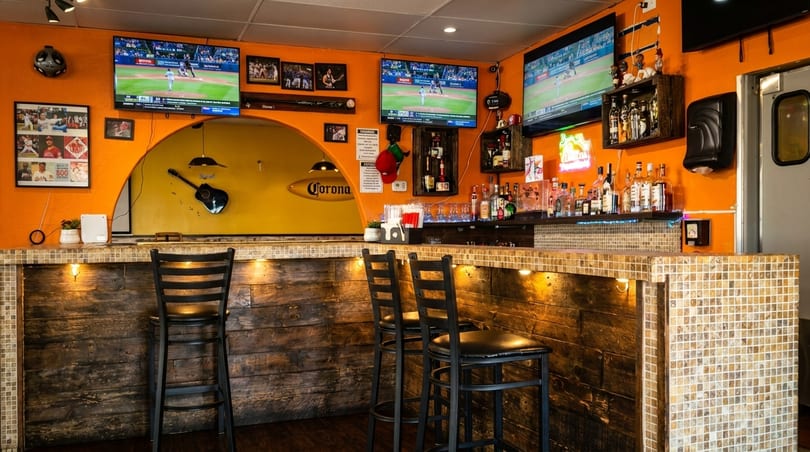 A vibrant bar with orange walls, multiple TVs, a tiled counter, and liquor bottles.